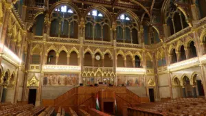 Wnętrze sali parlamentarnej z ozdobnymi drewnianymi ławami i bogatymi złotymi zdobieniami architektonicznymi.